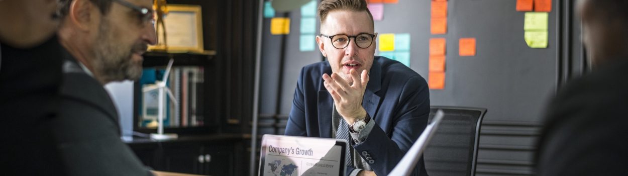 Homem de terno e óculos fala durante reunião em escritório moderno, sentado à mesa com laptop exibindo gráfico de crescimento da empresa, enquanto colegas escutam; ao fundo, parede com vários post-its coloridos organizados. Imagem de capa para o blogpost Como grandes corporações estão usando inovação aberta para acelerar decisões estratégicas