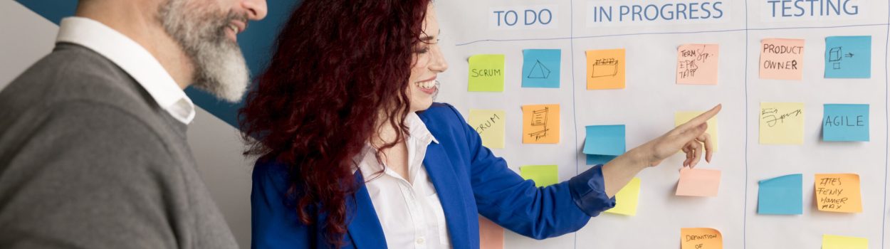 Um homem e uma mulher olhando para uma lousa cheia de post-it. A lousa está dividida em três colunas: A Fazer; Em Progresso, e Testando, com a mulher apontando para a coluna em progresso, para simbolizar práticas de startups