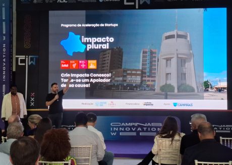 Painel de Led congelado no slide Impacto Plural, programa de aceleração de startups. No palco, à esquerda, estão o CEO da Venture Hub e a diretora executiva da Obará Edutech, responsáveis pelo programa.