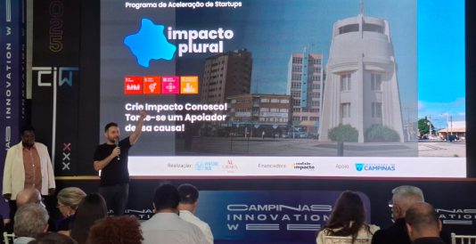 Painel de Led congelado no slide Impacto Plural, programa de aceleração de startups. No palco, à esquerda, estão o CEO da Venture Hub e a diretora executiva da Obará Edutech, responsáveis pelo programa.