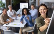 Equipe diversa em reunião de trabalho em um escritório moderno, enquanto uma mulher escreve em um quadro branco à frente do grupo e os colegas acompanham a discussão ao fundo. Imagem de capa para o blogpost: Como alinhar inovação aberta com estratégia corporativa sem criar “teatro de inovação”