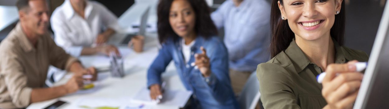 Equipe diversa em reunião de trabalho em um escritório moderno, enquanto uma mulher escreve em um quadro branco à frente do grupo e os colegas acompanham a discussão ao fundo. Imagem de capa para o blogpost: Como alinhar inovação aberta com estratégia corporativa sem criar “teatro de inovação”