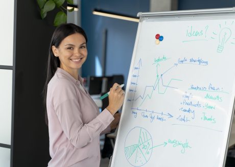 Mulher sorrindo em ambiente corporativo, escrevendo em um quadro branco com gráficos, anotações e ideias estratégicas durante uma apresentação ou reunião de negócios. Imagem de capa para o blogpost: A importância da inovação aberta em 2026.