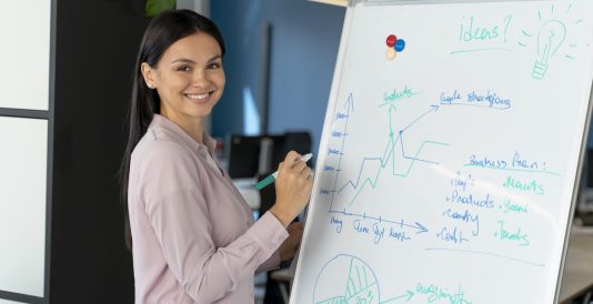 Mulher sorrindo em ambiente corporativo, escrevendo em um quadro branco com gráficos, anotações e ideias estratégicas durante uma apresentação ou reunião de negócios. Imagem de capa para o blogpost: A importância da inovação aberta em 2026.