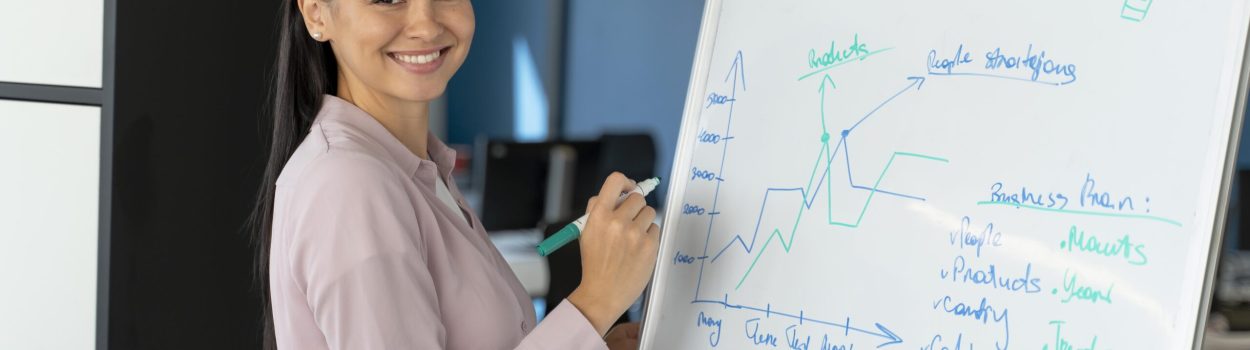 Mulher sorrindo em ambiente corporativo, escrevendo em um quadro branco com gráficos, anotações e ideias estratégicas durante uma apresentação ou reunião de negócios. Imagem de capa para o blogpost: A importância da inovação aberta em 2026.