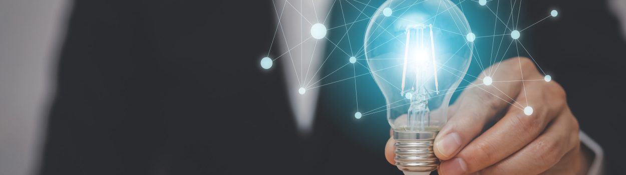 Pessoa de terno segurando uma lâmpada acesa com uma rede digital de conexões ao redor, acima de blocos de madeira com ícones de negócios, representando inovação, criatividade, tecnologia e estratégias empresariais. Imagem de capa para o blogpost: Inovação Aberta: Aproveitando Expertise Externa para Vantagem Competitiva