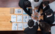 Vista superior de uma equipe de profissionais ao redor de uma mesa com relatórios e gráficos; eles posicionam engrenagens de madeira juntas, simbolizando colaboração, alinhamento estratégico e trabalho em equipe. Imagem de capa para o blogpost Da ideia à escala: o que muda quando a inovação aberta precisa rodar dentro da operação