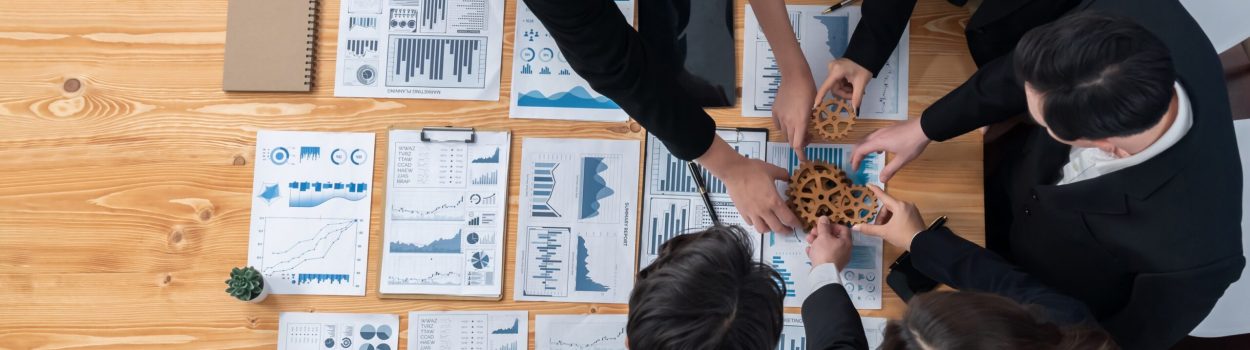 Vista superior de uma equipe de profissionais ao redor de uma mesa com relatórios e gráficos; eles posicionam engrenagens de madeira juntas, simbolizando colaboração, alinhamento estratégico e trabalho em equipe. Imagem de capa para o blogpost Da ideia à escala: o que muda quando a inovação aberta precisa rodar dentro da operação