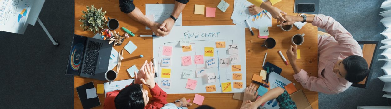 Vista superior de uma equipe diversa reunida em torno de uma mesa, colaborando na criação de um fluxo de trabalho com post-its coloridos, gráficos e anotações, representando planejamento estratégico, trabalho em equipe e metodologia ágil. Imagem de Capa para o blogpost Como Construir uma Cultura de Inovação